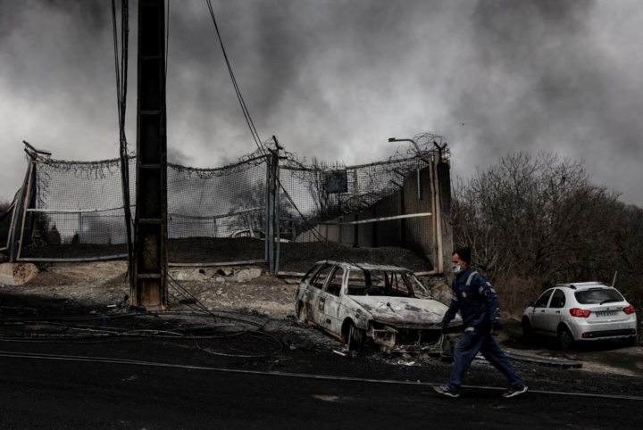 Người đàn ông đi ngang qua chiếc xe bị phá hủy khi khói bốc lên sau vụ tấn công nhằm vào các bồn nhiên liệu Shahran tại Tehran.