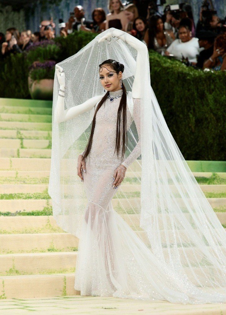 Lisa (BlackPink) trông như thiên thần ngoài đời thực trong chiếc váy trắng xuyên thấu lấp lánh tại Met Gala, được tô điểm thêm bằng chiếc khăn voan cầu kỳ. Thiết kế độc đáo mang theo cánh tay in 3D được dùng để nâng chiếc khăn voan cũng giúp Lisa trở thành tâm điểm chú ý.